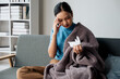 © aksonsat - Woman is ill sick with fever, a runny nose and cough feeling under weather under blanket in the living room at home and wipe a nose with facial tissue.