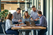 © SejutaCahaya - A team of five business professionals gathers around a large wooden table in a modern office, discussing ideas and looking at a laptop and papers.