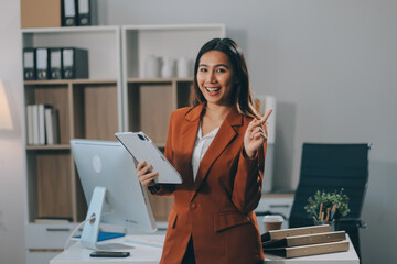 Attractive smiling Asian businesswoman standing holding tablet working and recording work details in office.