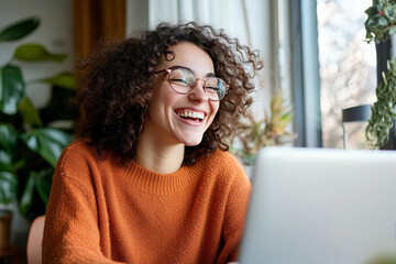 A young woman with curly hair and glasses laughs while looking at a laptop screen.