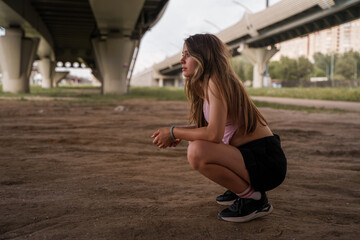 A stylish Caucasian young teenage girl is squatting under a bridge.