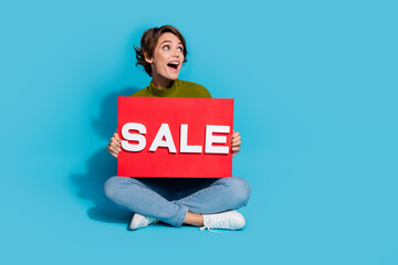 Poster - Photo of adorable cute cheerful girl sitting floor hands hold card look announcement empty space isolated on cyan color background