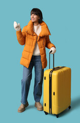 Wall Mural - Beautiful young happy woman with suitcase pointing at something on blue background. Winter vacation concept