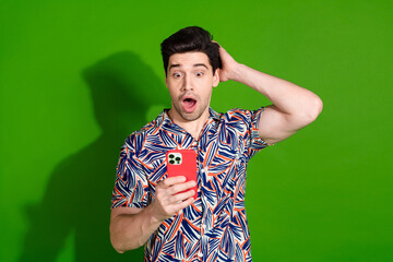 Photo of astonished guy with stylish haircut dressed print shirt staring at smartphone arm on head isolated on green color background