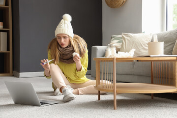 Wall Mural - Ill young woman with thermometer and laptop sitting at home