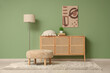 © Pixel-Shot - Chest of drawers with cushion, pouf, picture and floor lamp near green wall in room