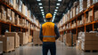 © khonkangrua - A warehouse worker in a safety vest and hard hat inspects a large storage aisle filled with boxes and stocked shelves.