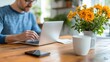 © LifeMedia - A man is focused on his laptop at a wooden table, adorned with a vase of yellow flowers, creating a balanced workspace with an inviting and productive atmosphere.