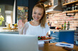 © Geber86 - Smiling woman eating salad while video chatting from home kitchen