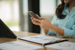 © Kritdanai - Businesswoman Checking Phone: A close-up image of a businesswoman's hands holding a smartphone, reviewing financial data on a tablet and clipboard, showcasing the seamless integration of technology in
