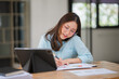 © Kritdanai - Focused & Connected: A young professional woman, in a stylish blue shirt, sits at her desk, engrossed in a phone call while taking notes on a tablet. Sunlight streams through the window, creating a wa