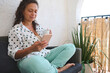 © Taras Grebinets - Relaxed woman sitting on a balcony with coffee, using smartphone for leisure or communication on a peaceful morning