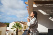 © Taras Grebinets - Woman enjoying morning coffee on sunny Mediterranean balcony with peaceful view of rooftops and palm trees.