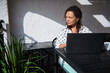 © Taras Grebinets - Woman working on laptop outdoors in sunlight at a patio table with a plant, wearing a polka dot blouse, enjoying a relaxed work environment.