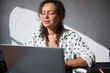 © Taras Grebinets - Woman working on laptop in a bright comfortable space with natural light and coffee, wearing a polka dot shirt, focusing on productivity and relaxation