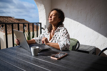 Woman enjoys sunlight on balcony while working on laptop, feeling relaxed and peaceful during a remote work day