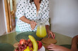 © Taras Grebinets - Woman in polka dot shirt serving fresh lemonade at a dining table with a bowl of assorted fruits including bananas, apples, and grapes