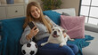 © Krakenimages.com - A young blonde caucasian woman enjoying her time indoors, sitting on a couch in her home with her pet canine, using a smartphone, and relaxing in the living room.
