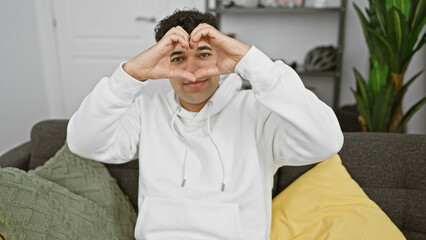 Wall Mural - A cheerful man gestures a heart with his hands, smiling in a cozy living room setting, showing positivity and comfort.