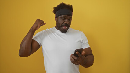 Handsome young african american man with a beard celebrating while looking at his smartphone against an isolated yellow background.