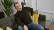 © Krakenimages.com - Hispanic man experiencing back pain while sitting on a gray couch in a modern living room.