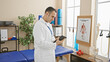 © Krakenimages.com - Hispanic man in white coat using tablet in rehab clinic room with exercise equipment and anatomy poster.