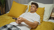 © Krakenimages.com - Young hispanic man relaxing on bed in a bedroom at home watching tv with a remote control in hand.