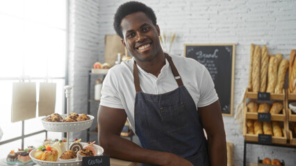 Handsome young african american man in a bakery shop wearing an apron and standing near an assortment of fresh pastries and bread