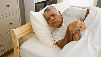 A middle-aged man lies in bed with eyes closed, appearing uneasy or unwell in a simple bedroom setting.