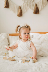 Wall Mural - Cute beautiful girl baby sitting on the bed in the bedroom and playing in eco wooden toys cubes and pyramid