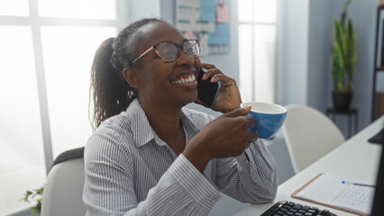 Wall Mural - Woman smiling while talking on phone and holding coffee in an office with natural light from large windows