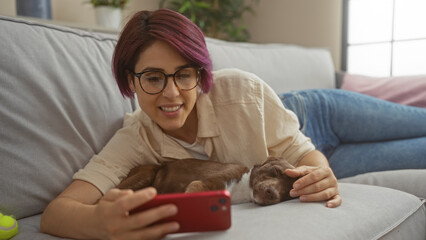 Poster - A young beautiful caucasian woman lies on a living room couch indoors, smiling at her phone while her pet dog sleeps beside her.