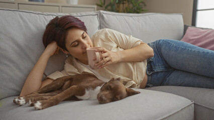 Poster - Young caucasian woman relaxing with her pet dog on a couch in the living room, sipping from a cup in a cozy home interior setting.