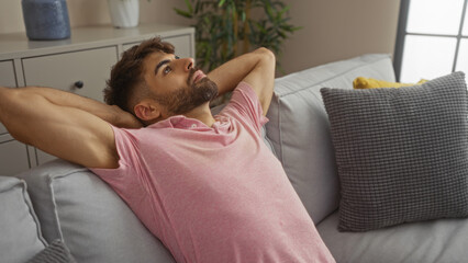 Young man relaxing on a sofa in a living room, wearing a pink shirt, and looking thoughtful, indoors, lying comfortably with hands behind head and pillows adding comfort to the scene