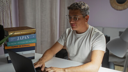 Young man working indoors on laptop at home with glasses, surrounded by books in a cozy living room setting