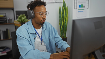 Young african american man with beard and glasses wearing headphones, working in an office at his computer in a modern workspace, focused and engaged.