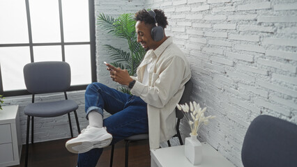 Young african american man with headphones smiling and using a phone while seated in a modern waiting room with chairs and indoor plants.