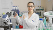 © Krakenimages.com - Young hispanic woman in glasses smiling and pointing in a laboratory setting, surrounded by scientific equipment.