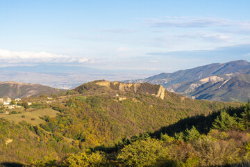 Hills covered with autumn trees illuminated by the evening sun. Part of the village is visible. Mountains and valleys in the background. Bright blue sky