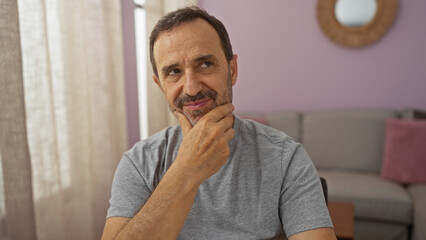 Sticker - Middle-aged hispanic man indoors sitting thoughtfully in a living room with light curtains and pink walls reflecting contemplation