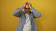 © Krakenimages.com - Hispanic male with glasses in a gray shirt expresses frustration by holding his head, standing against an isolated yellow background.