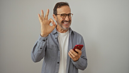 Poster - Middle-aged hispanic man holding a smartphone and showing an ok gesture isolated on white background