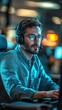 © Thann - Focused man in a headset working on a computer in a dimly lit room, possibly a tech support or customer service professional.