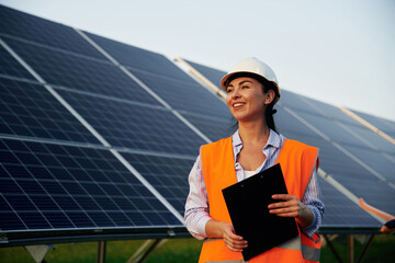 Black notepad in hands. Female worker engineer in uniform is near solar panels