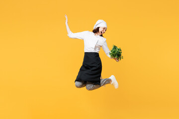 Full body young fun chef cook baker woman wear white shirt black apron uniform toque hat jump high hold in hand bunch of green parsley isolated on plain yellow background studio. Cooking food concept.