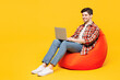 © ViDi Studio - Full body young IT man he wears red checkered shirt casual clothes sit in bag chair hold use work on laptop pc computer isolated on plain yellow orange background studio portrait. Lifestyle concept.