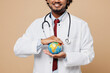 © ViDi Studio - Close up cropped young Indian doctor man wear white medical gown suit work in hospital clinic office hold in palm globe Earth map isolated on plain beige background studio Healthcare medicine concept