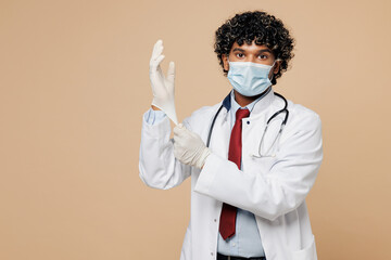 Wall Mural - Young confident Indian doctor man wear white medical gown suit mask work in hospital clinic office put on rubber gloves isolated on plain beige background studio portrait. Healthcare medicine concept.