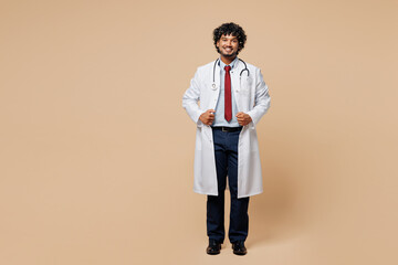 Wall Mural - Full body young smiling fun confident Indian doctor man wear white medical gown suit work in hospital clinic office look camera isolated on plain beige background studio. Healthcare medicine concept.