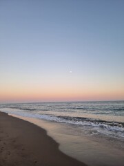 A serene beach scene in Marbella, Spain, captured at twilight with the moon rising over the horizon, and the soft colors of a Mediterranean sunset reflecting on the calm ocean waters.